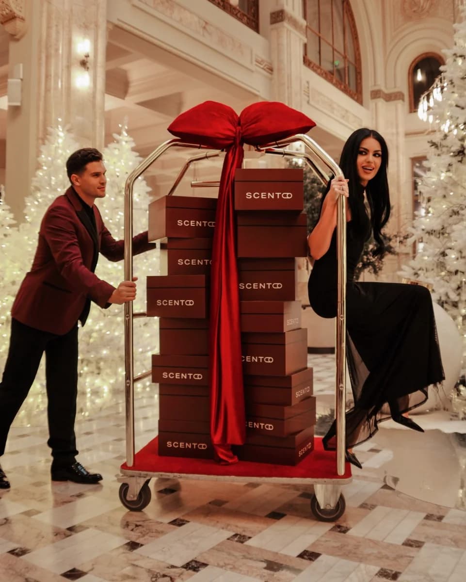 Ein Gepäckwagen im Hotel, beladen mit Scento-Geschenkboxen unter einer großen roten Schleife